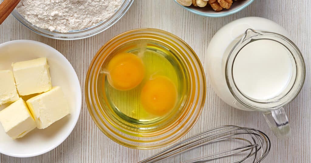 overhead view of bowl of raw eggs, a bowl of butter and a container of cream for baking