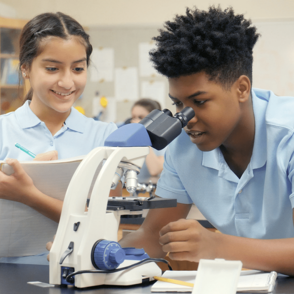 a teenage boy discusses his findings with his science lab partner as he looks into a microscope at their school
