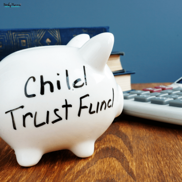 A white piggy bank labeled for a child trust fund sits on a wooden desk beside books and a calculator, symbolizing financial planning.