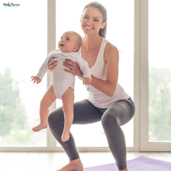 A smiling mother holding her baby while doing a squat exercise on a yoga mat at home.
