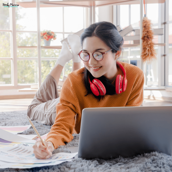 A teenager lying on the floor with headphones around her neck, working on a laptop and taking notes on colorful charts.