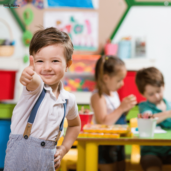 A smiling young boy giving a thumbs up in a colorful preschool classroom while other children engage in activities in the background.