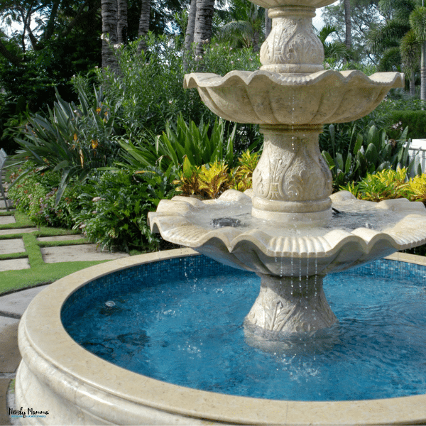 A tiered stone outdoor fountain with water flowing into a blue basin, surrounded by lush greenery and flowering plants.