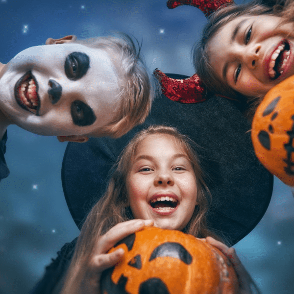 Three children dressed in Halloween costumes smile and hold carved pumpkins while looking down at the camera under a starry night sky.
