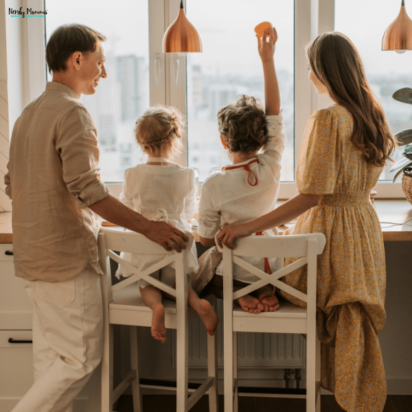 A family of four at home gathers at a kitchen counter by a large window, enjoying a bright morning together.