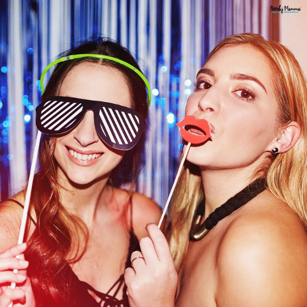 Two women smiling and posing with playful photo booth props at a celebration, representing fun entertainment ideas for planning a party with friends and family.
