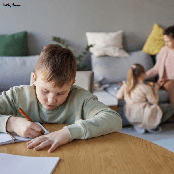 Child working at a table on school assignments while household activity continues behind him, reflecting practical ways to help children maintain attention during study time at home.