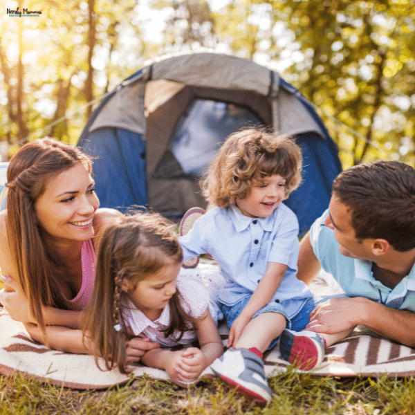 Family lying on a blanket in front of a tent outdoors, smiling and enjoying a relaxing camping trip together.