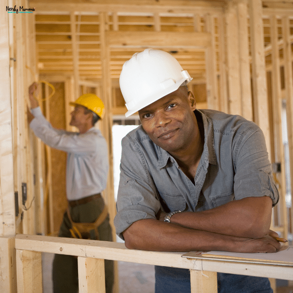 Professional renovation contractor wearing a safety helmet standing inside a home under construction while another worker installs wiring.