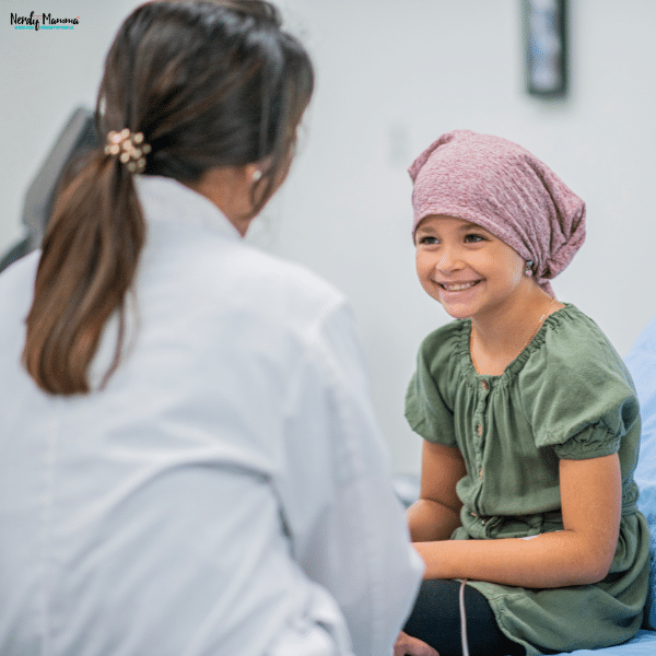 A young child wearing a headscarf smiles while sitting on an exam table and talking with a healthcare professional, showing comfort and support during mesothelioma care.