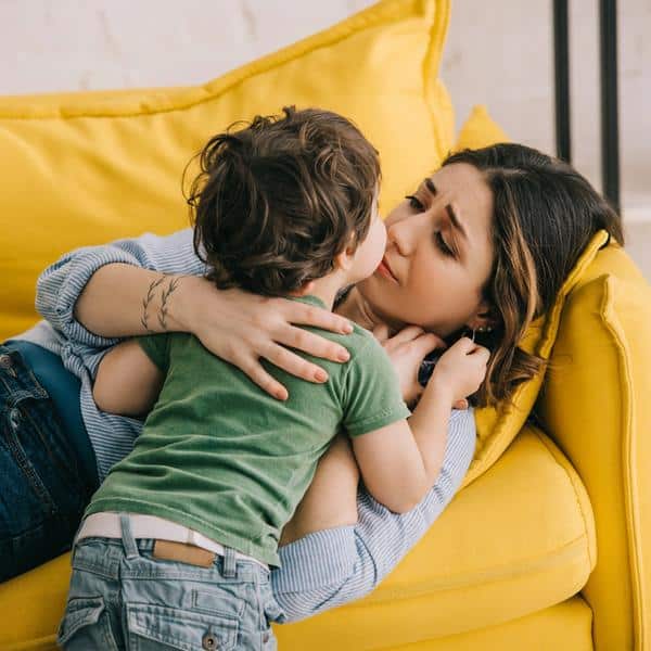Mother lying on a couch while comforting a young child during an emotional parenting moment that reflects stress, exhaustion, and mental load.