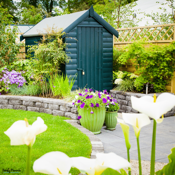 Small backyard garden shed surrounded by flowers, greenery, and landscaped pathways designed for outdoor storage and garden organization.