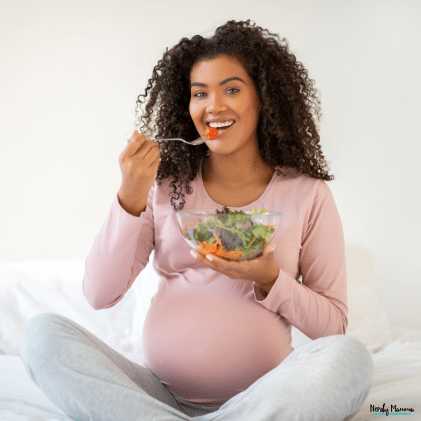 Pregnant woman sitting on a bed while eating a fresh salad, showing healthy pregnancy nutrition and balanced meal choices for moms-to-be.