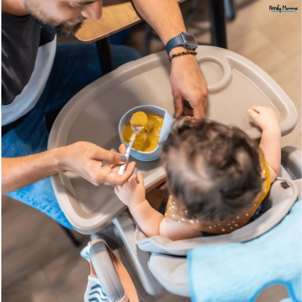 Parent feeding a toddler seated in a foldable high chair during mealtime at a family dining area.
