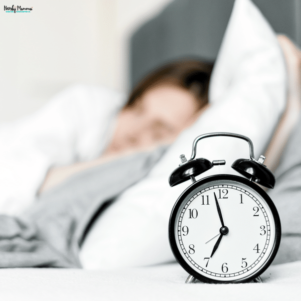 Alarm clock beside a bed with a sleeping person in the background, representing a consistent sleep routine for better daily energy.