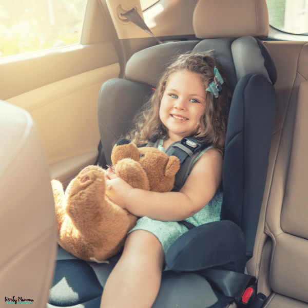 Smiling young girl rides securely in a properly installed booster car seat while holding a teddy bear during a family drive.