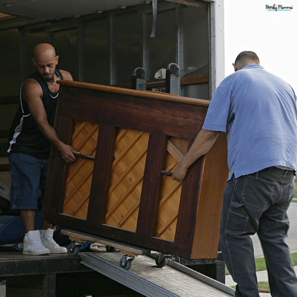 Two movers carefully loading an upright piano into a moving truck using a wheeled dolly and ramp, showing the equipment and labor involved in piano moving costs.