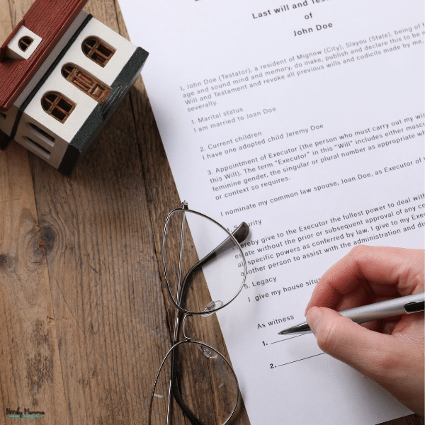 Hand signing a will beside a model house and eyeglasses, representing estate planning for property, assets, and a family’s future.