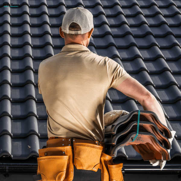 A professional roofing contractor wearing a tool belt installs dark roof tiles while carrying a stack of replacement materials.