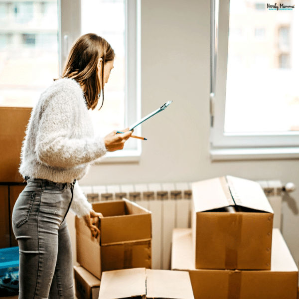 A woman reviews a moving checklist on a clipboard beside cardboard boxes in a partly packed room.