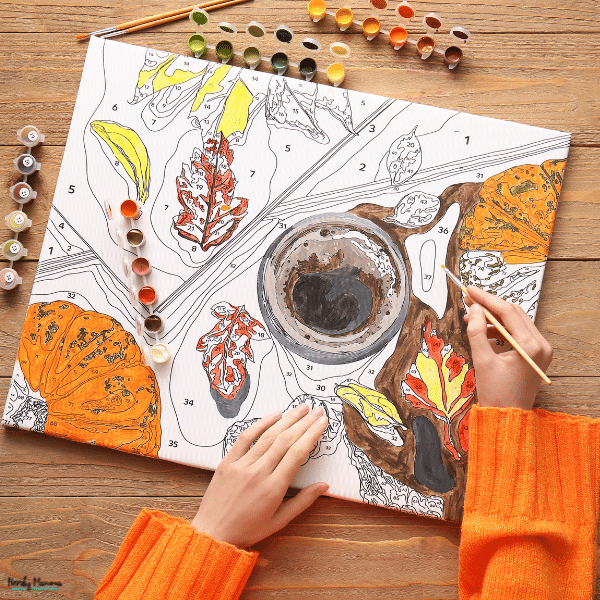 A paint-by-numbers kit includes a fall-themed numbered canvas, colorful paint pots, and brushes as a person fills in the design.