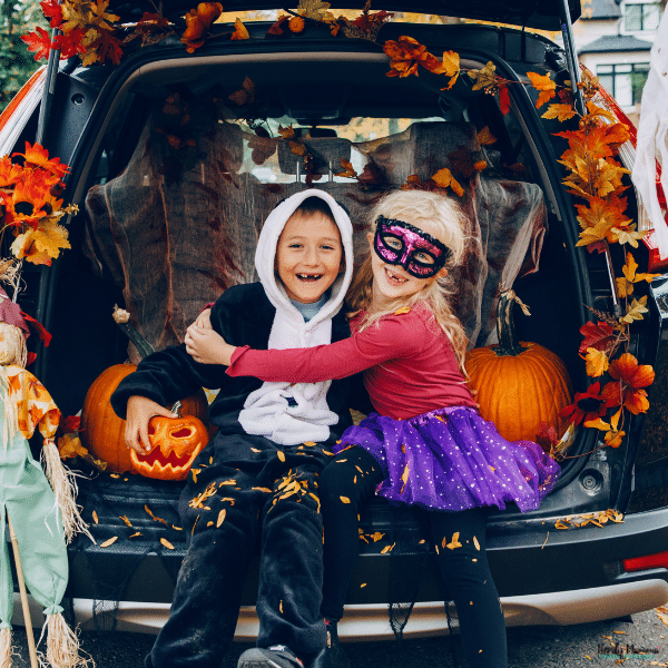 Two costumed children sit in a pumpkin-filled car trunk decorated for a trunk-or-treat event.