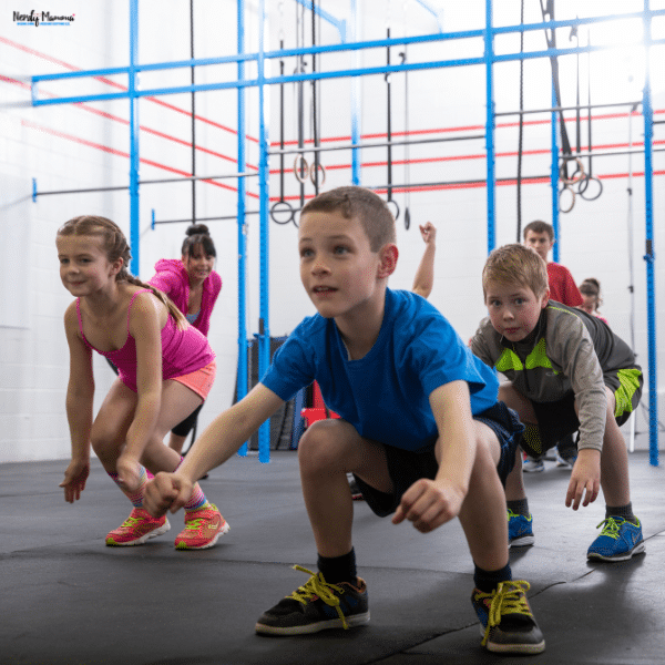 Children practice squats in an indoor fitness space equipped with climbing frames and hanging rings.
