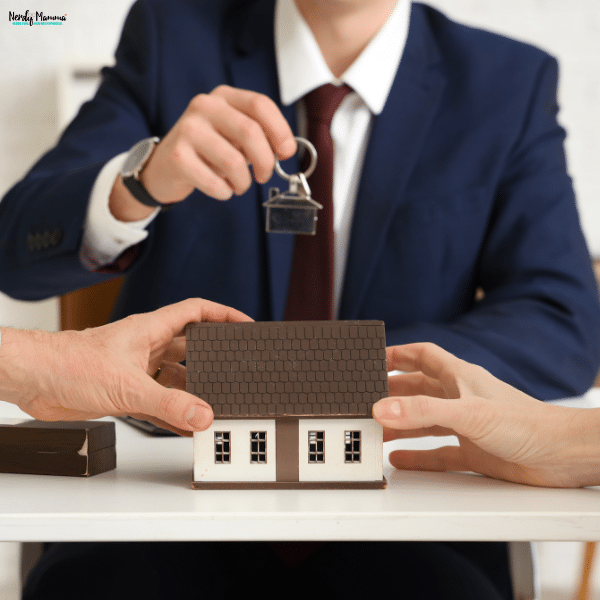 A suited professional holds house keys above a model home between two people, illustrating home ownership discussions in marital property settlements.