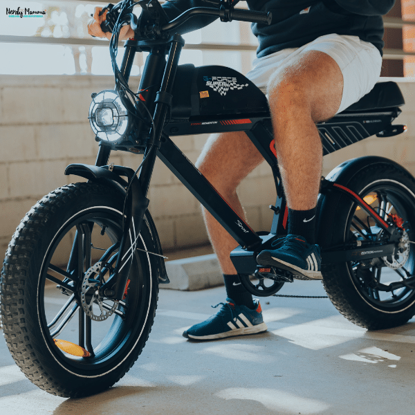 Person in white shorts sits on a black fat-tire e-bike with a large front headlight and one foot on a pedal.
