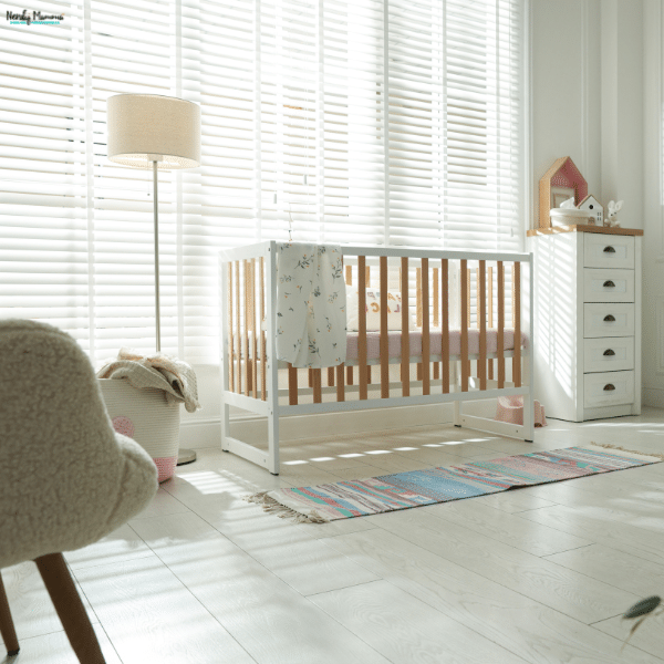 White horizontal nursery blinds filter sunlight behind a wooden crib, with a floor lamp and narrow dresser nearby.
