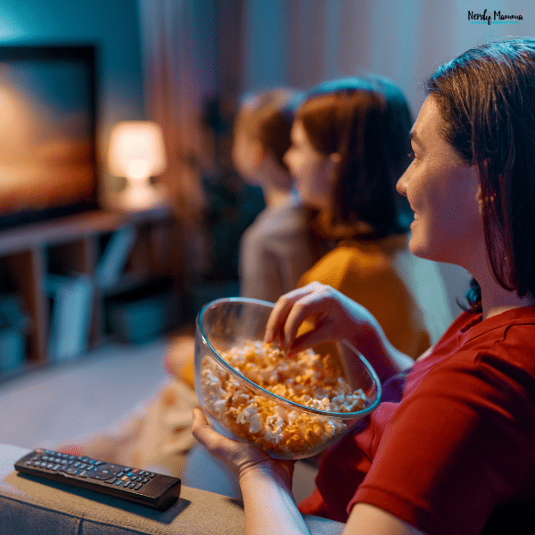 A woman holds a bowl of popcorn while watching television with two companions during a cozy fall rewatch night.