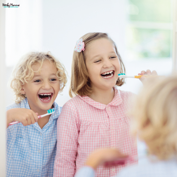 Two smiling children practice dental hygiene with colorful toothbrushes while looking in a bathroom mirror.