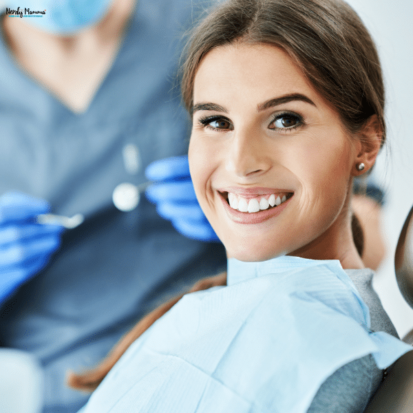 A smiling patient reclines in a treatment chair while a masked dentistry professional holds instruments behind her.