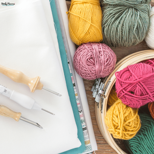 Punch needle tools rest on folded fabric beside wooden embroidery hoops and colorful balls of yarn.
