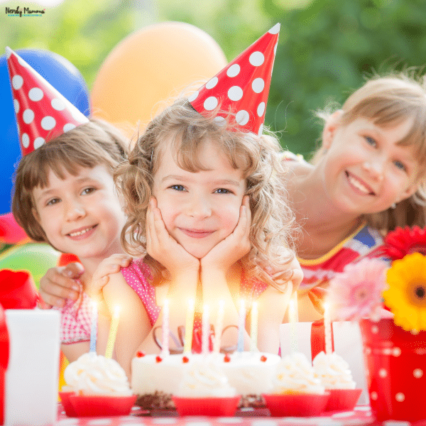 Three smiling children celebrate a kids birthday party outdoors with a candlelit cake, cupcakes, and colorful balloons.