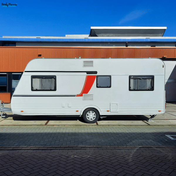 White travel trailer with curtained windows, a red accent stripe, and lowered stabilizer legs parked outside an orange building under a clear blue sky.