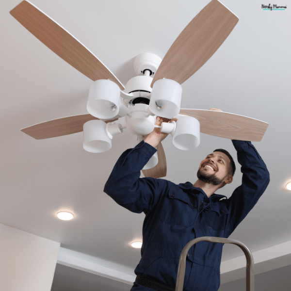A man on a stepladder installs a white ceiling fan with wooden blades and four light shades.