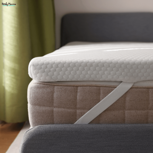 A white mattress topper with a textured cover sits on a beige mattress, held in place by an elastic corner strap.
